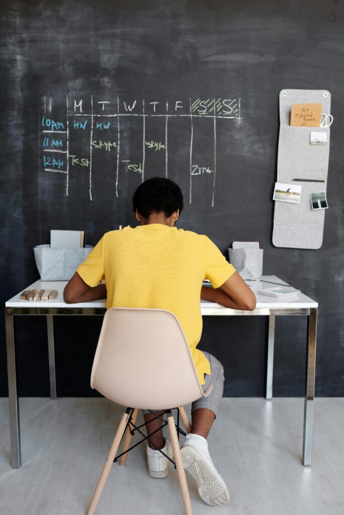 pexels-photo-4144101-4144101 A teenager focuses on studying at a desk, with a blackboard displaying a schedule in the background.